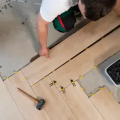 Pose d'un carrelage imitation bois sur un sol en béton. Un ouvrier ajuste les lames, des cales jaunes et noires sont visibles.