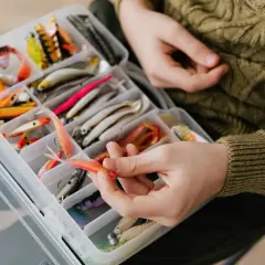 Ręka wybiera pomarańczową przynętę z pudełka pełnego różnorodnych akcesoriów do łowienia na żywca.