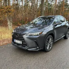 Ciemnoszary Lexus NX450h+ na mokrej drodze w jesiennym lesie.