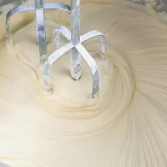 Mieszadła miksera obracają się w gładkim, kremowym **kremie z mascarpone i białej czekolady**, tworząc wiry.