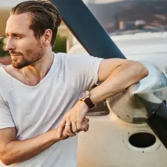 Mężczyzna w białej koszulce opiera się o samolot, zastanawiając się, jak być bardziej atrakcyjnym. Jego spojrzenie skierowane jest w dal, a na nadgarstku ma elegancki zegarek.