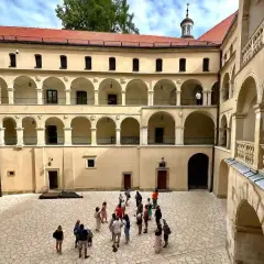 Dziedziniec zamku z arkadowymi krużgankami, kamienna posadzka przypomina pieskowa skała. Grupa turystów zwiedza historyczne miejsce.