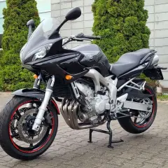 Czarny motocykl Yamaha FZ6, gotowy do jazdy, z czerwonymi akcentami na felgach, stoi przed budynkiem z zielonymi drzewami.