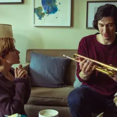 Adam Driver håller i en trumpet i en scen från en av hans filmer. Bredvid honom sitter en kvinna och tittar på.