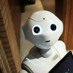 Der humanoide Roboter Pepper mit leuchtenden Augen und blauen Akzenten hält ein Tablet. Er ist bereit für eine audio ki-gesteuerte Interaktion.