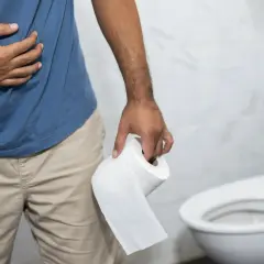 Mężczyzna trzymający papier toaletowy, z ręką na brzuchu, w łazience. Prawdopodobnie doświadcza biegunki po alkoholu.
