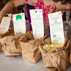 Barn plockar popcorn ur papperspåsar. En påse med popcorn med kolasås syns tydligt.