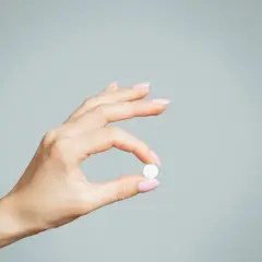 Dłoń z różowymi paznokciami trzyma białą tabletkę, być może zolpidem, na jasnoszarym tle.