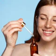 Uśmiechnięta kobieta z piegami aplikuje serum z pipetki. W buteleczce znajduje się tokoferol, znany ze swoich właściwości pielęgnacyjnych.