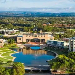 Weite Luftaufnahme des Campus der Bond University mit einem zentralen See, gepflegten Grünflächen und modernen Gebäuden, umgeben von Bäumen und Hügeln.