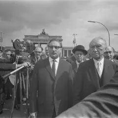 Männer in Anzügen, eine Filmkamera und das Brandenburger Tor im Hintergrund. Ein Mann hält eine "Stalin-Note" in der Hand.