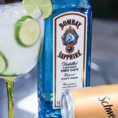 Orzeźwiający drink z tonikiem i limonką, obok butelka ginu Bombay Sapphire i puszka Schweppes. Idealny na letnie wieczory.