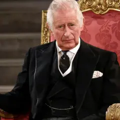 Król Karol w wieku siedemdziesięciu lat, w eleganckim czarnym garniturze, siedzi na ozdobnym tronie.