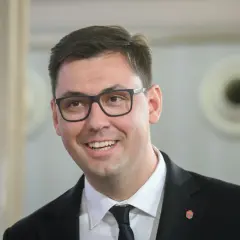 Uśmiechnięty Konrad Frysztak w okularach, w garniturze. Jego wykształcenie pozwala mu na pewność siebie w każdej sytuacji.