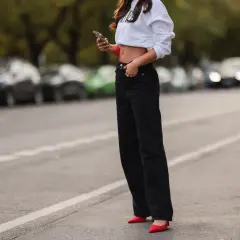Kobieta w białej bluzie i czarnych jeansach, z czerwoną szpilką na stopie, trzyma telefon.
