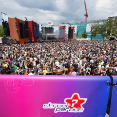 Folla immensa al festival techno di Zurigo, Street Parade, con DJ set e musica elettronica sotto un cielo nuvoloso.