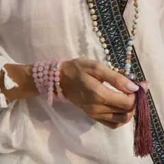 Mano con varios tipos de japa mala: uno de cuarzo rosa en la muñeca y otro largo con cuentas de colores y borla.