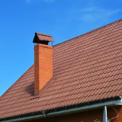 Ceglany komin z metalowym daszkiem na brązowym, falistym dachu. Miękka obróbka komina zapewnia mu estetyczny wygląd.