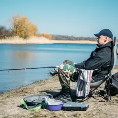 Mężczyzna siedzi na krześle nad jeziorem, łowiąc ryby metodą. Wędka zarzucona, obok torba i pudełko na akcesoria.