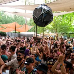 Folla entusiasta in un festival techno all'aperto, sotto tende e alberi. Luci e musica creano un'atmosfera vibrante.