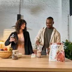 Ein Mann und eine Frau bereiten einen Proteinshake zu. Die Packung zeigt, wie wird Proteinpulver hergestellt.
