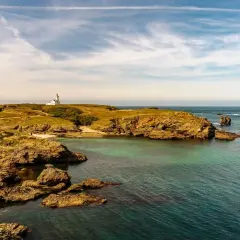 Phare blanc sur une colline verdoyante d'une île bretonne, avec des rochers dorés et une mer turquoise sous un ciel nuageux.
