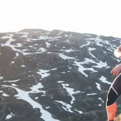 Une femme en tenue de sport s'assoit sur des rochers, regardant sa montre, prête à trouver un trail dans un paysage montagneux enneigé.