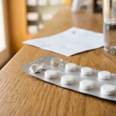 Opakowanie tabletek, w tym akamprozat, obok szklanki wody i recepty na drewnianym stole.