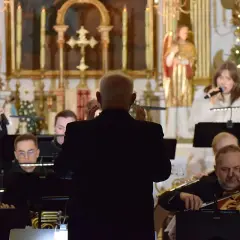 Dyrygent prowadzi orkiestrę i wokalistki w kościele, w tle choinka.