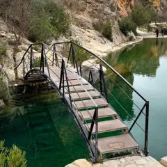 Puente metálico sobre aguas cristalinas, ideal para bolbaite fotos. Rocas y vegetación rodean el estanque.