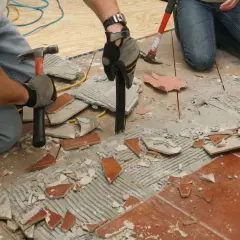 Deux personnes s'y prennent pour comment décoller du carrelage au sol, utilisant marteaux et burins pour briser les carreaux et le ciment.