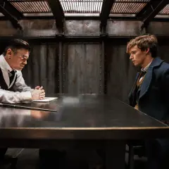 Scena z filmu "Fantastyczne zwierzęta i jak je znaleźć". Newt Scamander i Percival Graves siedzą przy stole, podczas gdy dwie kobiety stoją w tle.