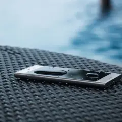 Telefon z kroplami wody na ekranie, leżący na plecionej powierzchni przy basenie. Klasa szczelności **IP 65** zapewnia ochronę przed zachlapaniem.