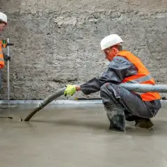 Dwóch budowlańców w kaskach i kamizelkach wylewa pianobeton z węża na podłogę. Jeden z nich kontroluje poziom.