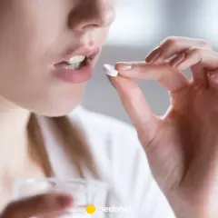 Kobieta bierze tabletkę clindamycin, trzymając ją między palcami. Obok widać szklankę z wodą.
