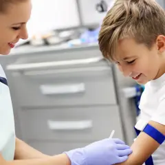 Uśmiechnięty chłopiec przechodzi badanie krwi. Pielęgniarka pobiera próbkę, by sprawdzić, czy nie ma podwyższonych płytek krwi u dziecka.