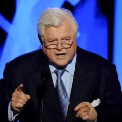 Ein älterer Mann mit Brille und grauem Haar spricht in ein Mikrofon. Er trägt einen dunklen Anzug mit einer blauen Krawatte. Seine Hand ist erhoben, als würde er eine Rede halten, die an die Kennedy-Krankheit erinnert.