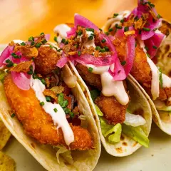 Tres deliciosos tacos de pescado crujientes, cubiertos con cebolla morada encurtida, salsa cremosa y cilantro fresco.