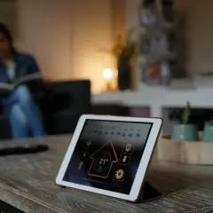 Tablette affichant l'interface d'un thermostat connecté pour chaudière gaz. Une femme lit sur un canapé en arrière-plan.