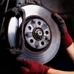 Mechanik w czerwonych rękawicach wymienia tarczę hamulcową. Hamulec ręczny trzyma jedno koło, zapewniając bezpieczeństwo podczas prac.