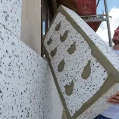 Budowlaniec montuje płytę z polistyrenu na ścianie. Materiał izolacyjny ma szary granulat.