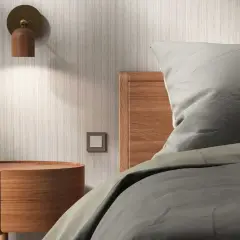 Lámpara de madera en la pared ilumina una mesita redonda y la cabecera de una cama con sábanas grises.