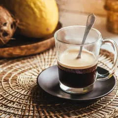 Tazza di caffè fumante su sottobicchiere intrecciato. Sullo sfondo, cocco e melone. Un caffè lungo ha più caffeina per iniziare la giornata.