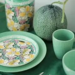 Assiette en bone china porcelaine ornée de fleurs colorées, à côté d'une tasse et d'un bol verts, avec un melon et un vase fleuri.