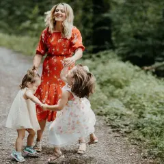 Mama w czerwonej sukience śmieje się, trzymając za ręce dwie córeczki. Dzieci wirują w radosnym tańcu, tworząc ciekawe zabawy taneczne na leśnej ścieżce.