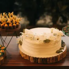 Biały tort komunijny ozdobiony białymi kwiatami i zielonymi liśćmi. Obok słodkości, które mogą być inspiracją, jakie kwiaty na tort komunijny.