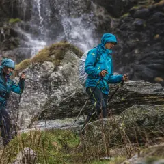 Dwie osoby w turkusowych kurtkach przeciwdeszczowych idą z kijkami trekkingowymi obok wodospadu.