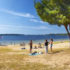 Słoneczna plaża nad jeziorem goczałkowickim. Ludzie odpoczywają, kąpią się i pływają na żaglówkach. Widoczna wieża ratownicza.