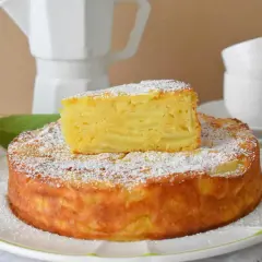 Pyszna torta di mele, posypana cukrem pudrem, z kawałkiem jabłka na wierzchu. Obok cytryna i jabłko.