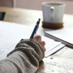 Dłoń w swetrze pisze długopisem, tworząc rzeczowniki odczasownikowe na papierze. Obok kubek kawy.